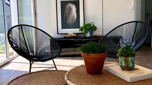 A modern living room with a large window, two black wireframe chairs, a wicker table, a floor lamp, a framed artwork, and a potted plant. - The Saint Vaast orchard  (Saint-Vaast-la-Hougue)