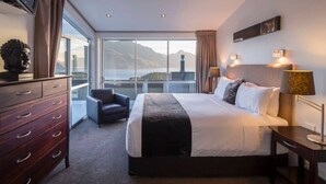 A hotel room with a large bed, a dresser, a chair, and a view of mountains and a body of water. - Alpen View Villa | Short walk to town (Queenstown)
