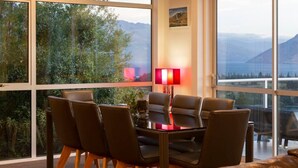 A dining area with a table and chairs, large windows with a mountain view, and a red lamp on the table. - Alpen View Villa | Short walk to town (Queenstown)