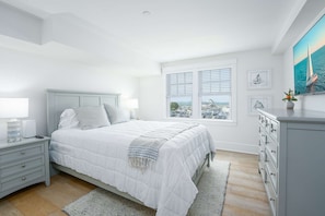 Un dormitorio con cama, mesita de noche, cómoda y vista al mar a través de la ventana.