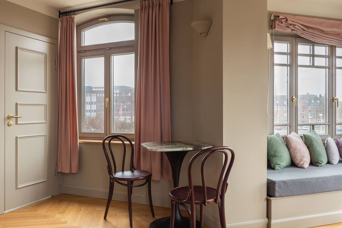 A room with a bay window, two chairs, a small table, and a cushioned bench with pillows.