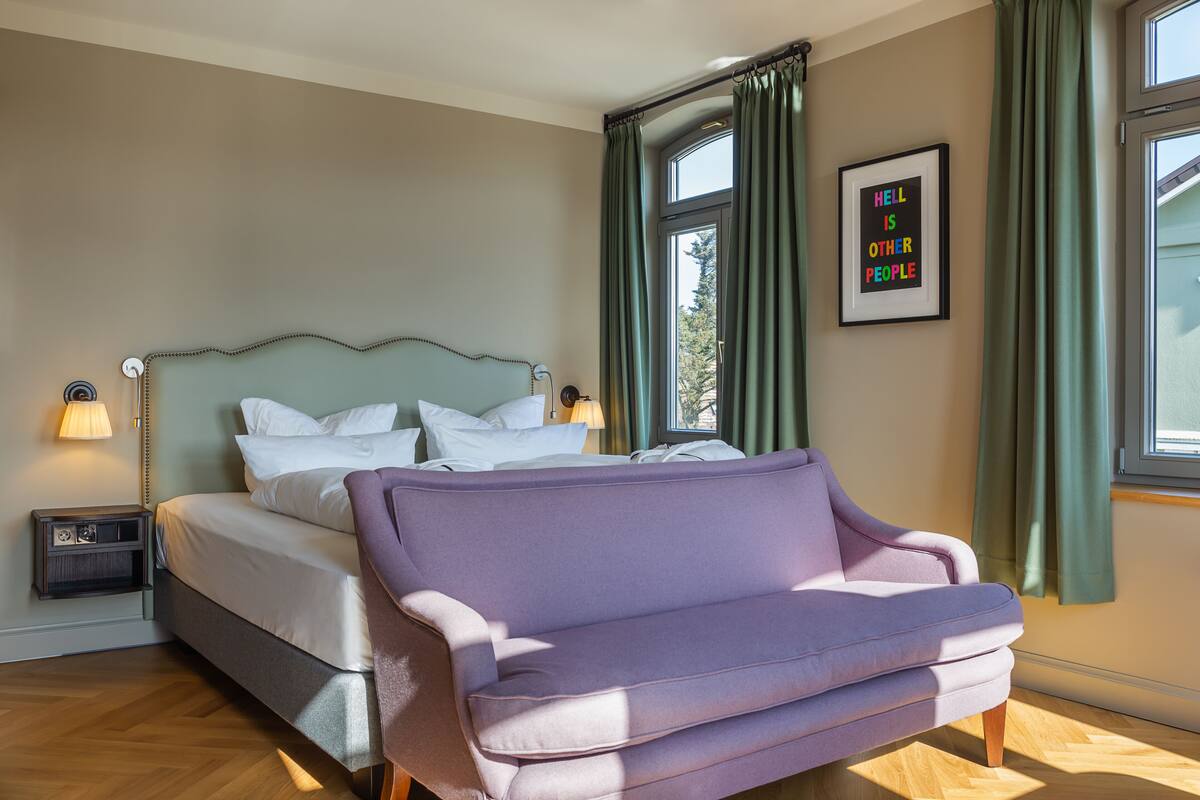 A bedroom with a bed, a purple armchair, a nightstand, a lamp, a window with curtains, and a framed quote on the wall.