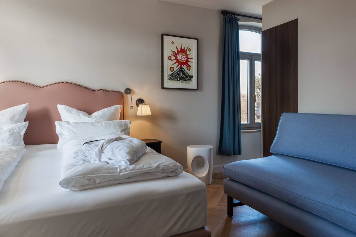 A hotel room with a bed, a bedside table with a lamp, a framed picture on the wall, a window with curtains, and a blue sofa.