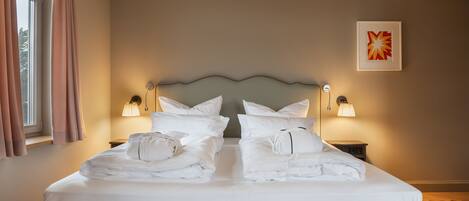 A neatly made bed with pillows and a folded towel, bedside lamps, a framed artwork on the wall, and a window with curtains.