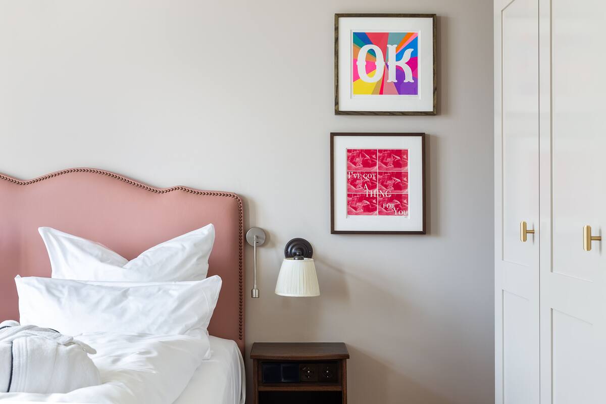 A neatly made bed with white linens and pillows, a bedside table with a lamp, and two framed artworks on the wall.