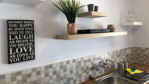 Cocina con paredes blancas, salpicadero de azulejos y encimera de madera. Hay tres repisas sobre el fregadero, cada una con varios objetos.