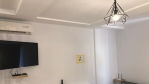 Una sala de estar moderna con un televisor de pantalla plana, un estante empotrado con una botella y aperitivos, un sofá gris con un cojín a rayas y una lámpara colgante geométrica.