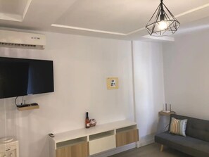 A modern living room with a flat-screen TV, a built-in shelf with a bottle and snacks, a grey sofa with a striped pillow, and a geometric pendant light.