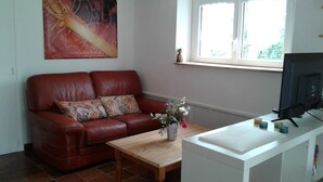 A living room with a red leather sofa, a wooden coffee table, a white cabinet with a television, and a window with a view of greenery. - Appartement au Calme Dans un Parc Ombragé (Montignac)