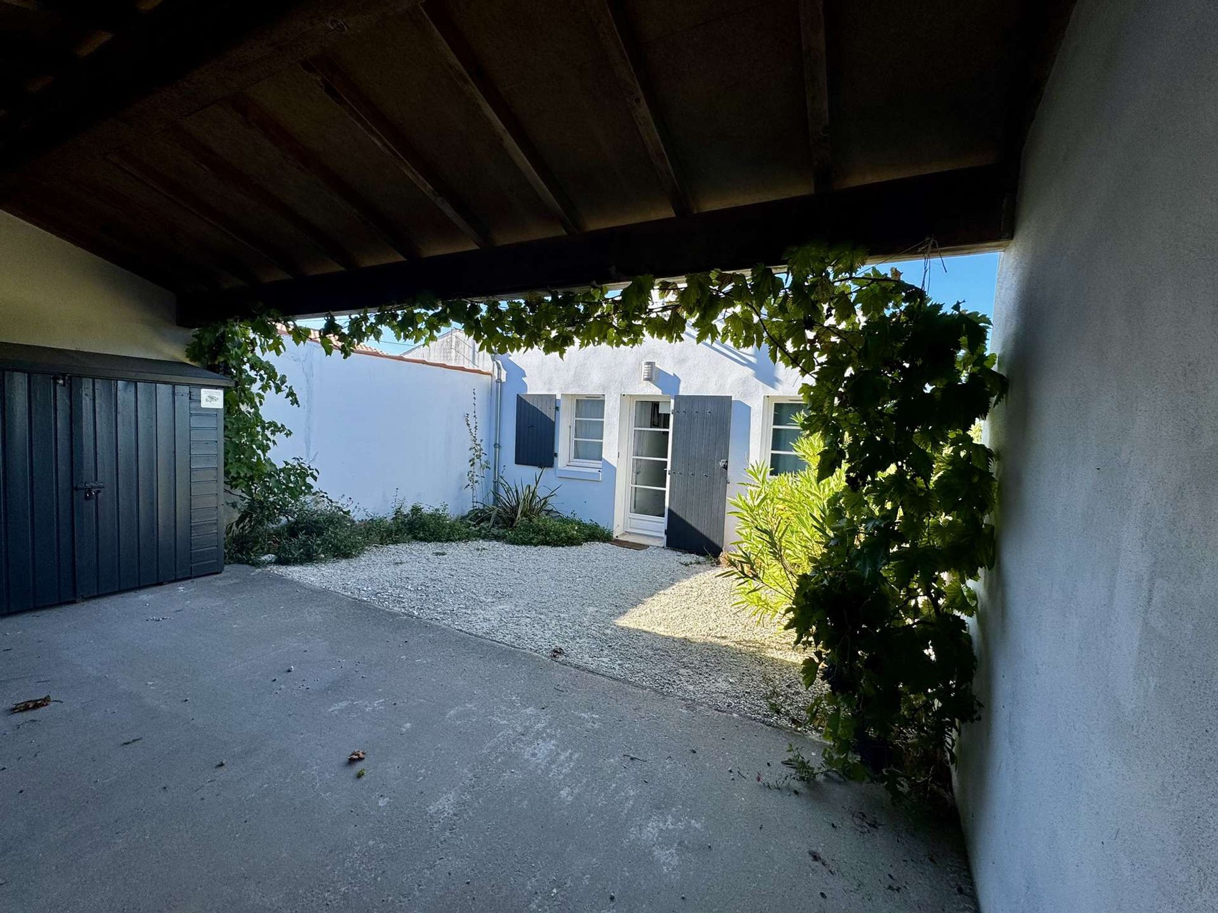 A covered outdoor area with a gravel path, a grey gate, and a building with a white facade and a window with a shutter.