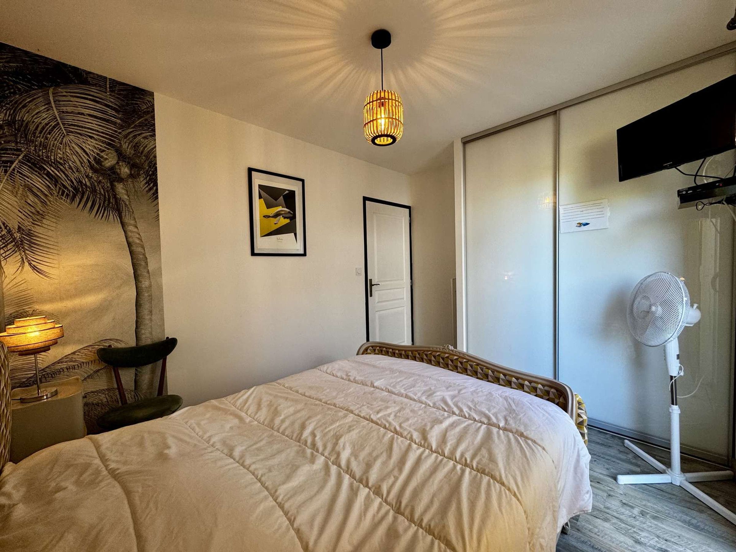 A bedroom with a bed, a wall-mounted TV, a ceiling light, a door, and a fan.
