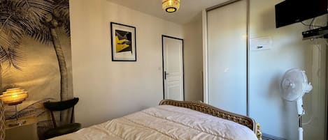 Une chambre à coucher avec un lit, un téléviseur fixé au mur, un luminaire au plafond, une porte et un ventilateur.