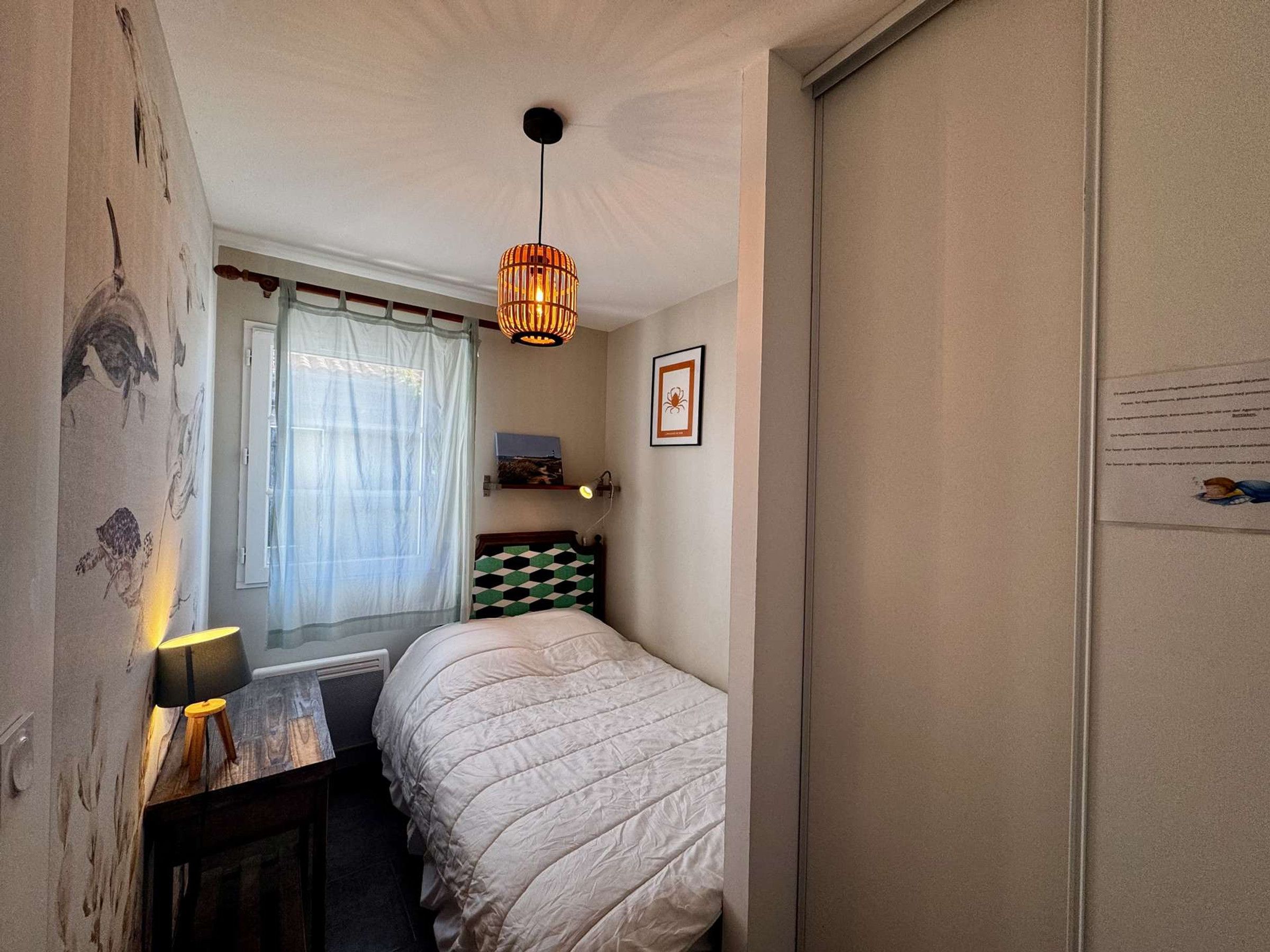 A bedroom with a bed, a bedside table with a lamp, a hanging light fixture, a window with curtains, and a wall with a framed picture.