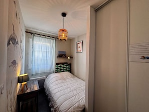 Un dormitorio con cama, mesita de noche con lámpara, lámpara colgante, ventana con cortinas y una pared con un cuadro enmarcado.