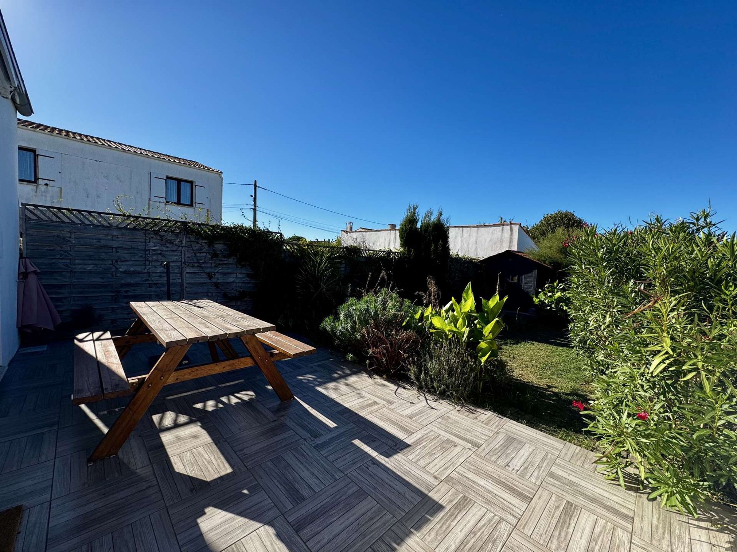 A wooden deck with a picnic table, surrounded by a garden with various plants and a stone wall.