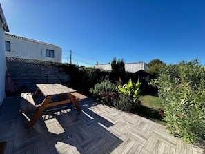 Une terrasse en bois avec une table de pique-nique, entourée d’un jardin où poussent différentes espèces de plantes et bordée d’un mur en pierre.