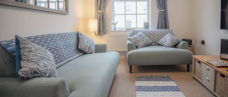 Un salon comprenant un canapé, un fauteuil plus petit, une table d’appoint avec une lampe, un téléviseur et une fenêtre avec des rideaux.