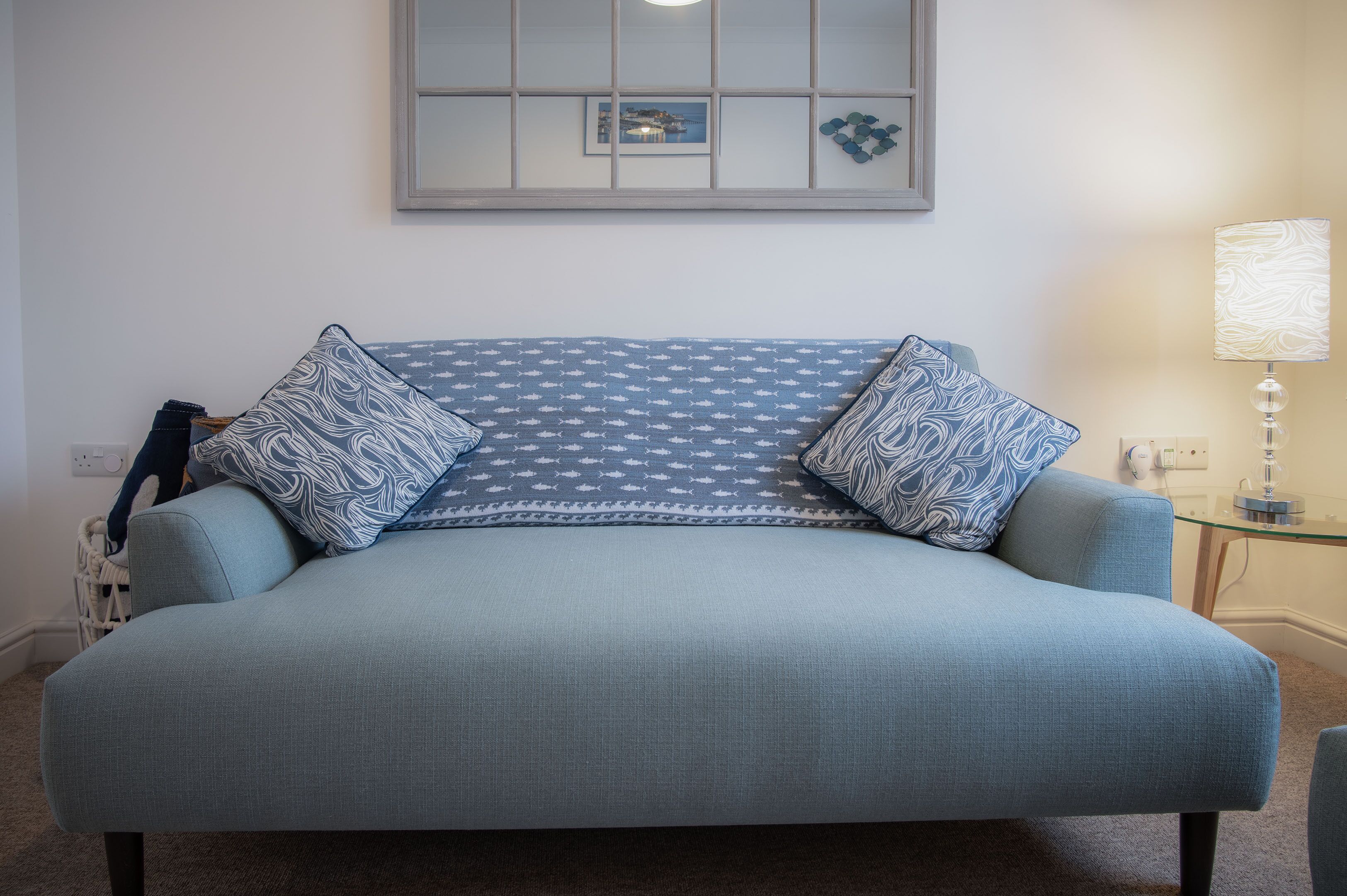 A light blue sofa with patterned cushions, a glass side table, a lamp, and a framed picture on the wall.