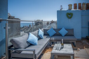 Un balcón con un sofá gris, cojines azules y una mesa con una botella y copas.