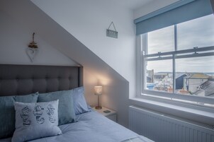 Un dormitorio con techo inclinado, una cama con ropa de cama gris, una mesita de noche con una lámpara y una ventana con vistas a edificios.