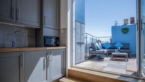 Una cocina moderna con armarios grises, un microondas y un lavamanos.  Al exterior, hay un balcón con un sofá y una mesa, y una pared azul decorada con un corazón.