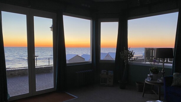 Ein Zimmer mit großen Fenstern und Blick auf einen Strand bei Sonnenuntergang, ein kleiner Tisch mit einer Lampe und ein Topfpflanze.