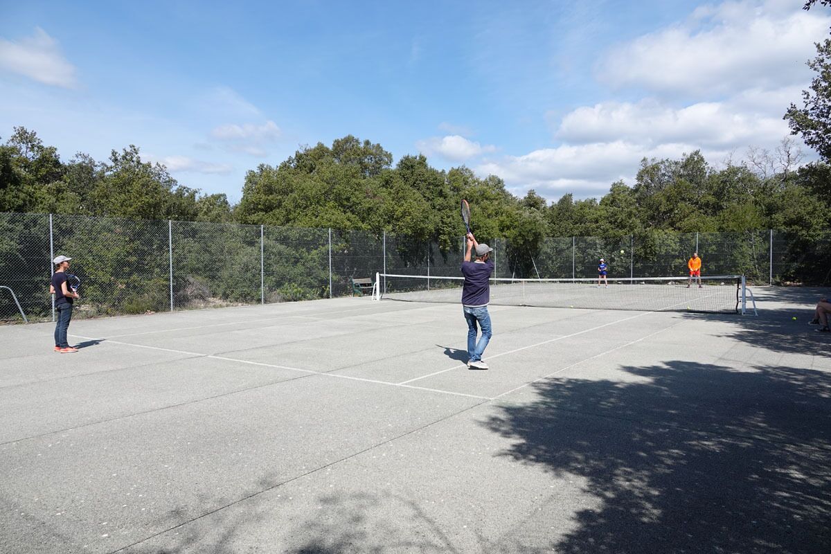 Un court de tennis en plein air, doté d’un filet, entouré d’une clôture en treillis métallique et d’arbres.