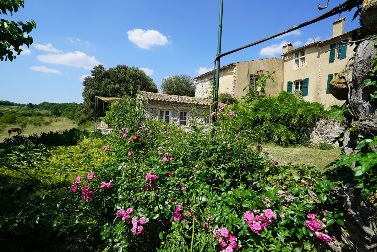 A rustic stone house with a garden in front, featuring pink flowers and a clear blue sky.