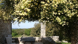 A stone patio with a table and chairs, surrounded by lush greenery and a stone wall. - La Grange Domaine de la Blaque (Varages)