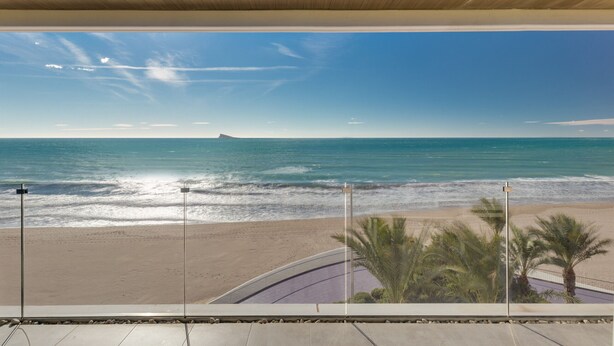 Une vue sur la plage depuis un balcon doté d’un plafond en bois, avec des palmiers et un ciel dégagé.