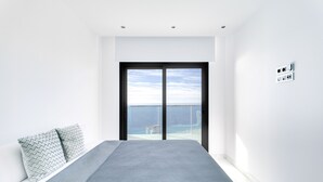 Une chambre moderne avec un grand lit, une vue sur la mer par une porte coulissante et un mur blanc doté d’une prise de courant.