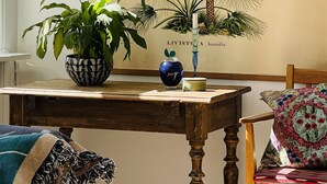 Ett rum med ett träbord, en hängande lampa av rotting, en krukväxt, en vas och en inramad botanisk illustration på väggen.