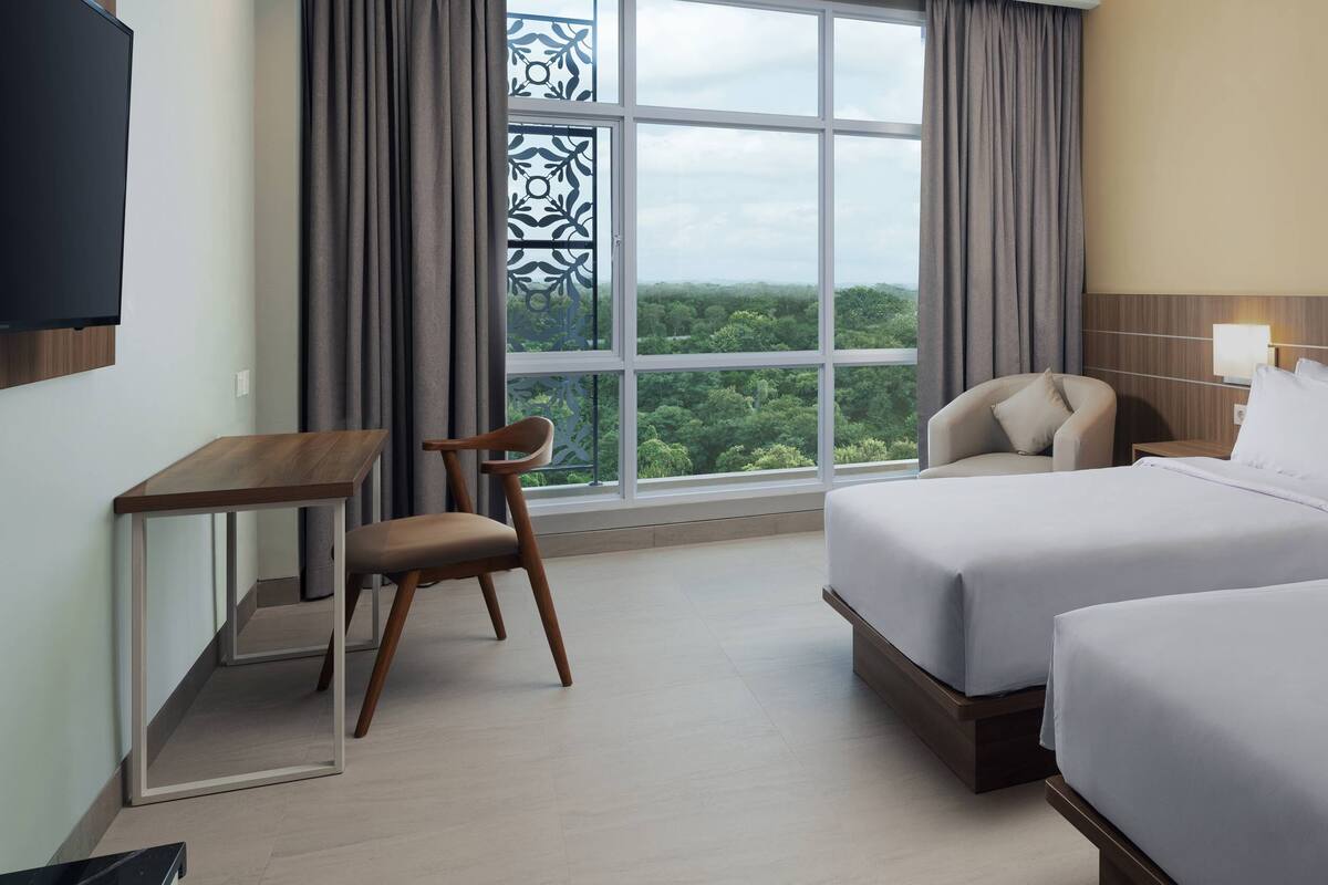 Ein Hotelzimmer mit zwei Betten, einem Schreibtisch, einem Stuhl und einem großen Fenster mit Blick ins Freie.