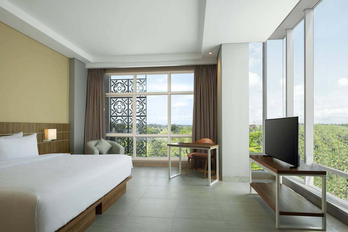 Ein Hotelzimmer mit einem großen Bett, einem Schreibtisch mit Fernseher und Blick auf Grün hinter großen Fenstern.