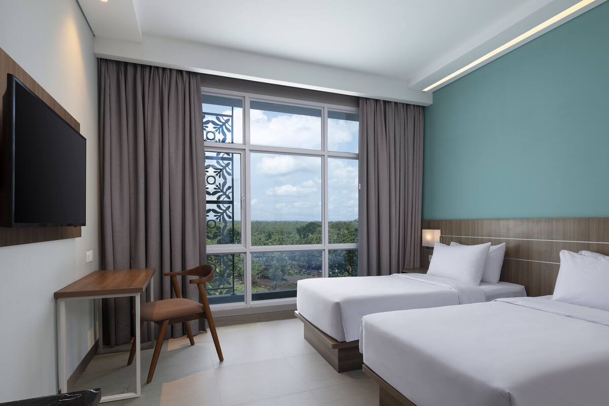 Ein Hotelzimmer mit zwei Betten, einem Schreibtisch, einem Stuhl, einem Fernseher und einem großen Fenster mit Blick auf Grünflächen.