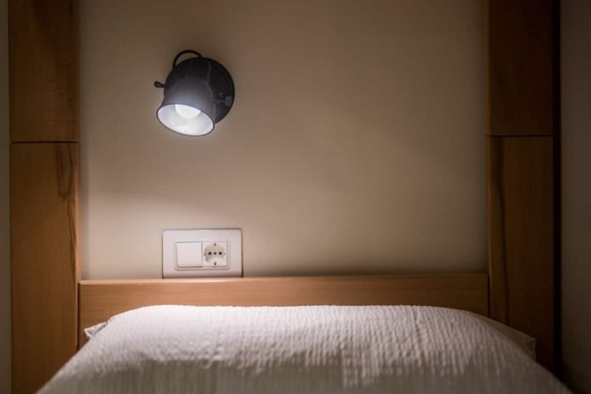 A single bed with a white mattress and pillows, a bedside table with a lamp, and a wall socket.