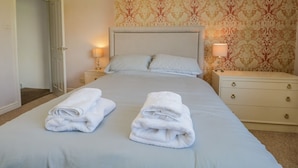 A neatly made bed with two folded white towels, a nightstand with a lamp, and a patterned wallpaper on the wall. - Viewpoint (Kendal)
