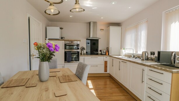 A modern kitchen with white cabinets, a wooden dining table, and a vase of flowers. - Viewpoint (Kendal)