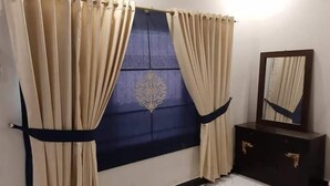 A room with a large window covered by beige curtains, a dark blue window shade, and a mirror on a dark wooden dresser. - Hotel Royal Defence (Lahore)