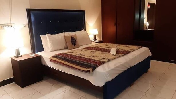 A hotel room with a large bed, two bedside tables, a nightstand, a headboard, and a wardrobe. - Hotel Royal Defence (Lahore)