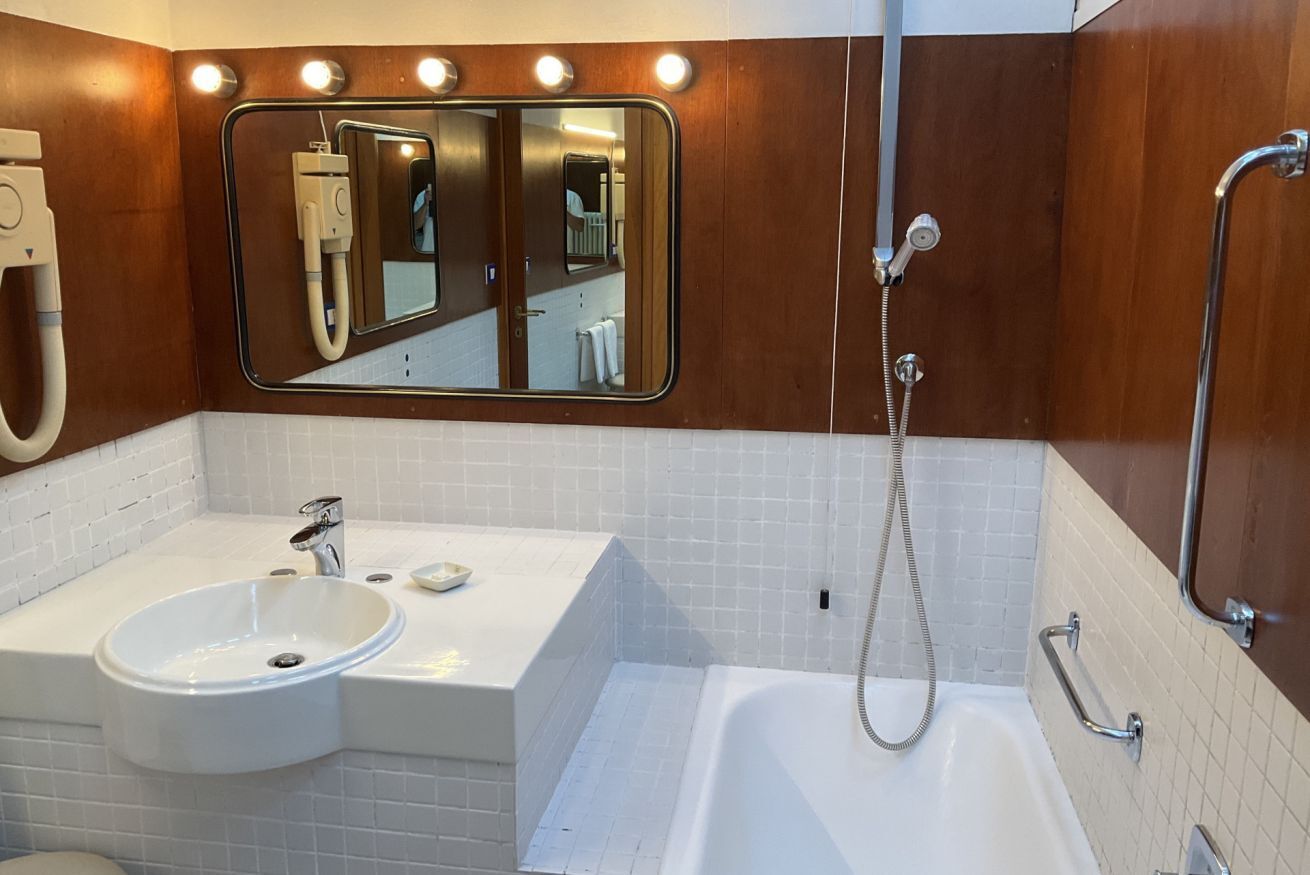 A modern bathroom with a white sink, a large mirror, a shower with a handheld hose, and wooden paneling.