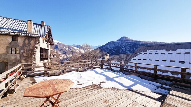 Una terraza de madera con una mesa, suelo cubierto de nieve y vistas a la montaña.