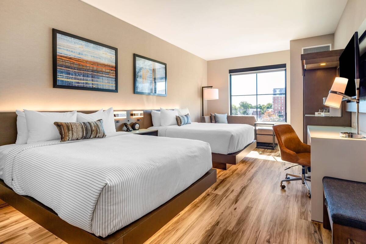A hotel room with two beds, a desk, a chair, and a large window.
