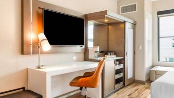 A modern hotel room with a white desk, a brown leather chair, a flat-screen TV mounted on the wall, a small kitchenette, and a large window providing natural light.