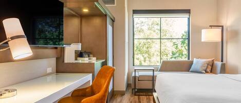 A modern hotel room with a desk, a comfortable chair, a bed, and a large window offering a view of trees.