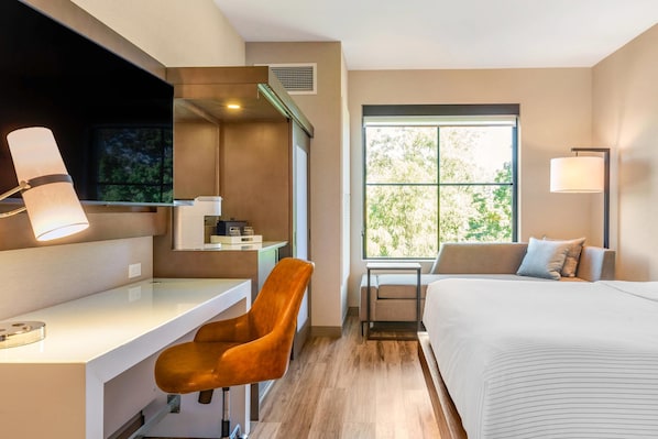Quarto de hotel moderno com escrivaninha, poltrona confortável, cama e uma grande janela com vista para árvores.