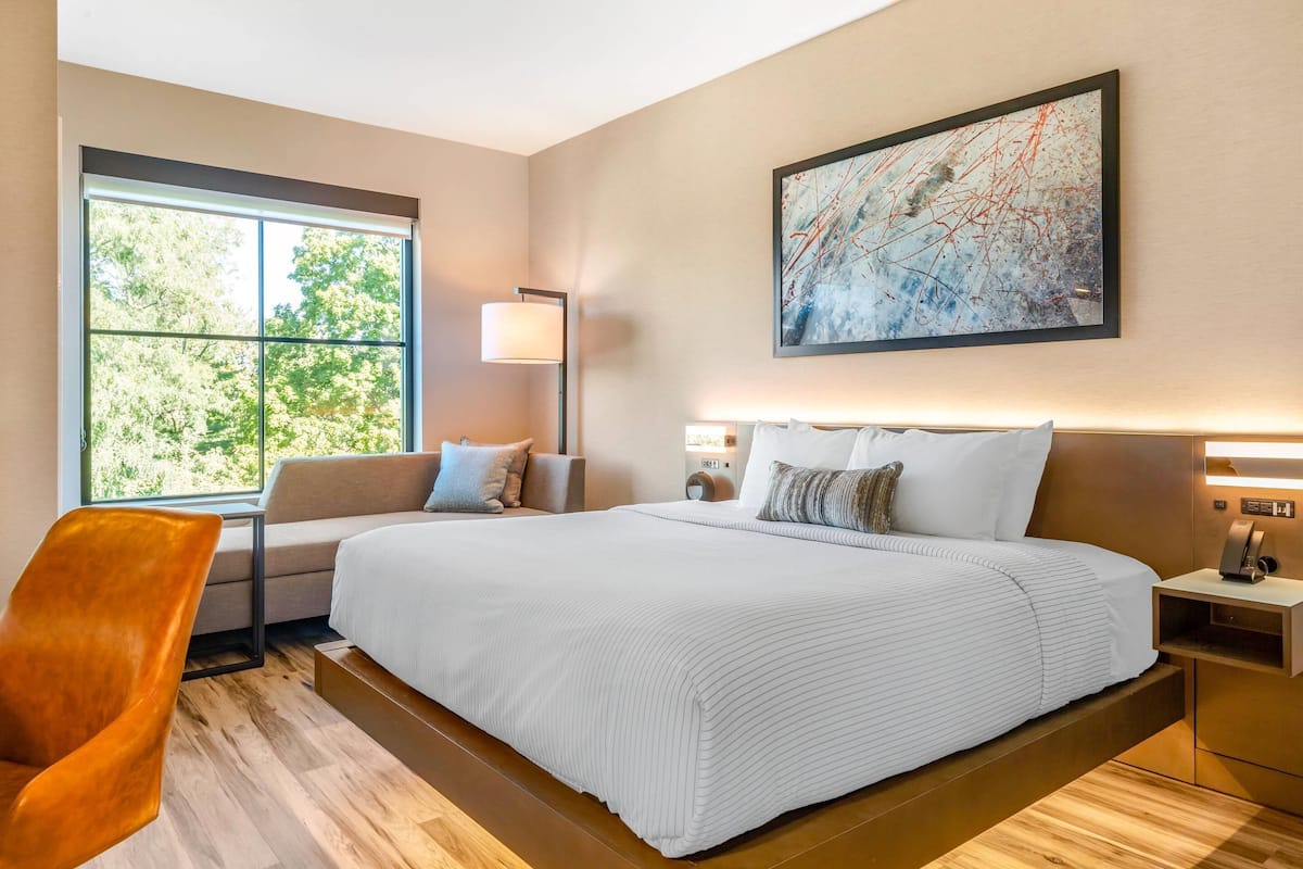 A modern hotel room with a large bed, a seating area with a sofa and armchair, a floor lamp, and a large abstract painting on the wall.