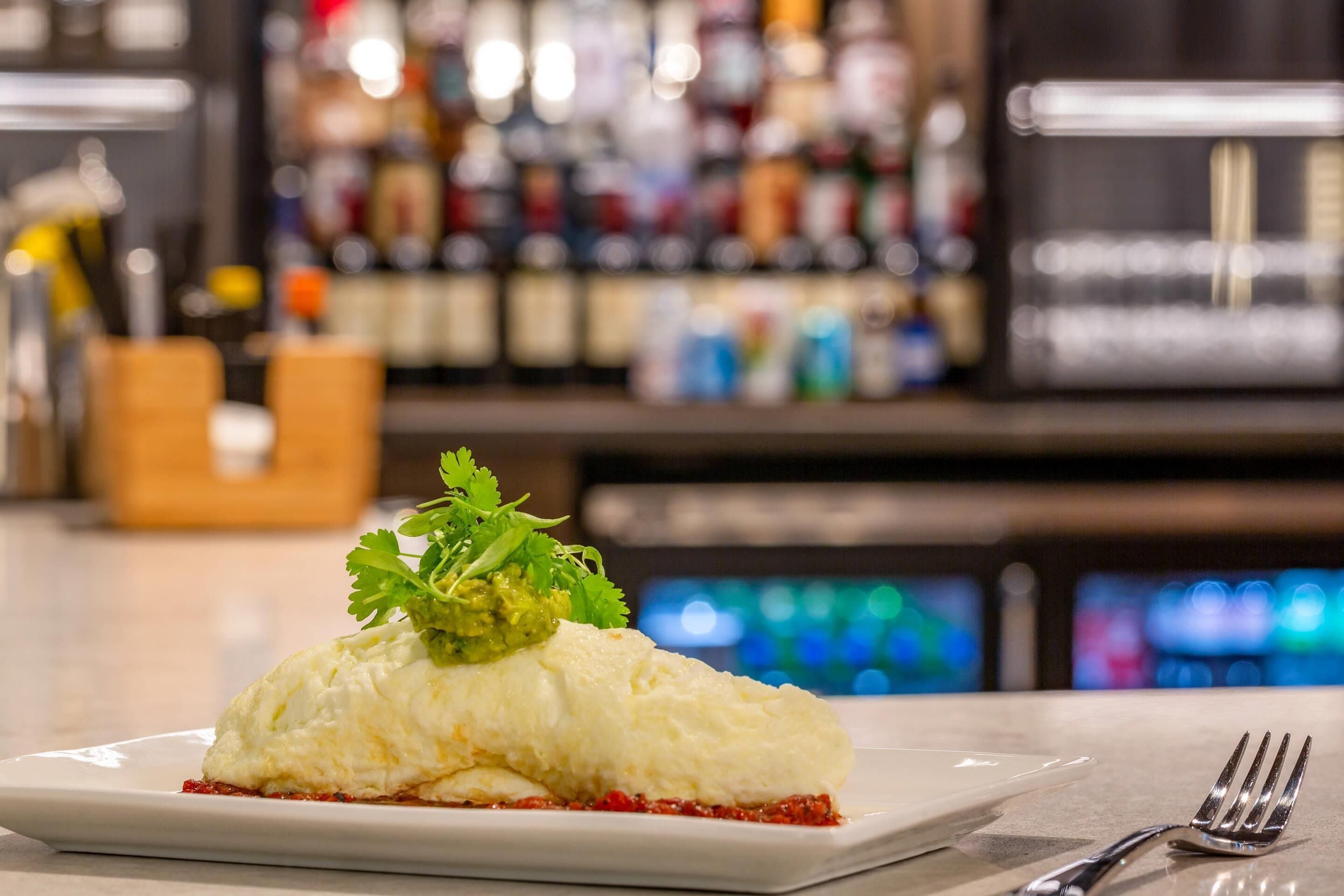 A plate with a food item, a fork, and a bar with bottles in the background.