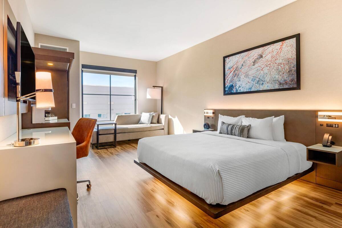A modern hotel room with a large bed, a desk, a chair, and a sofa. There is a large window and a painting on the wall.
