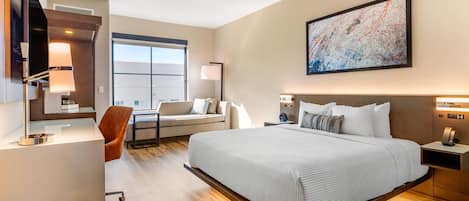 A modern hotel room with a large bed, a desk, a chair, and a sofa. There is a large window and a painting on the wall.